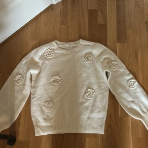 Vit stickad tröja med blommor Dobber - Superfin vit stickad tröja från Dobber i storlek S. Tröjan har långa ärmar och är dekorerad med söta virkade blommor framtill och på ärmarna. Perfekt för dig som gillar en söt och trendig look med unika detaljer.
