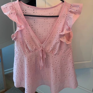 Ljusrosa blus med volangärmar och knyt - Supersöt ljusrosa blus med broderade hålmönster, volangärmar och v-ringning med knyt framtill. Luftig och flowy passform, perfekt för varma dagar. Ger en romantisk vibe och är lätt att styla med jeans eller kjol.
