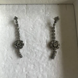 Glittriga silverörhängen med stenar - Säljer ett par snygga örhängen i silverfärg med glittrande stenar. De har en böjd form och en rund blomliknande detalj i mitten, täckt av små gnistrande stenar