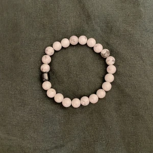 Vitt pärlarmband med marmormönster - Stilrent armband med runda vita pärlor i marmormönster och en svart pärldetalj. Elastiskt band gör det enkelt att ta på och av. Perfekt accessoar för en clean och modern look.