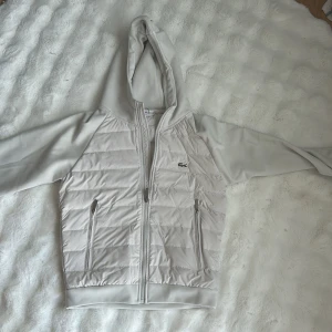 Vit Cardigan från Lacoste - Snygg vit cardigan från Lacoste med huva och dragkedja. Jackan har quiltad framsida och rygg, två dragkedjefickor och klassisk krokodillogga på bröstet. Perfekt för dig som vill ha en stilren och sportig look. 