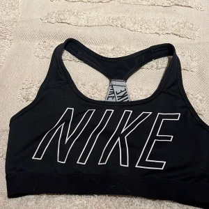 Svart sporttopp från Nike - Svart sporttopp från Nike med stort vitt logotryck framtill och korsade band, tight passform, perfekt för träning eller gymmet. Snygg och sportig design.