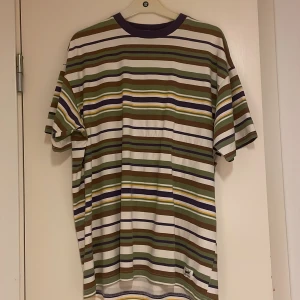 Randig t-shirt från Pull&Bear - Snygg randig t-shirt från Pull&Bear i storlek M. T-shirten har breda och smala ränder i vitt, grönt, brunt, lila och gult. Klassisk passform med korta ärmar och rund halsringning i lila. Perfekt för dig som gillar streetstyle och färg.