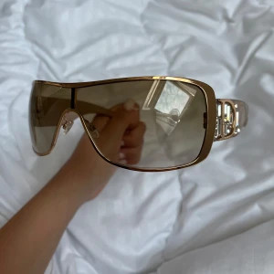 Äkta Dior indinight2 solglasögon. Champagne/guld  - Säljer ett par statement-solglasögon från Dior, modell Indinight2. De har stora, bruna shield-linser och en champagnefärgad ram med metallic-look. Sidorna är dekorerade med glittriga stenar och Dior-logga. Perfekt accessoar för att sticka ut och ge en lyxig vibe . Ursprungligt pris runt 4 000 kr – slutsåld modell. Pris går att diskutera. 