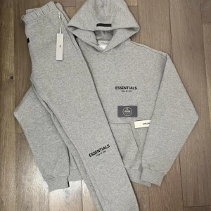 Söker en essientials track suit - Snygg grå hoodie från Essentials Fear of God med svart logga på bröstet. Klassisk modell med huva och ribbade muddar. Perfekt för en chill och avslappnad stil. Mjuk bomullsblandning som känns skön mot huden.