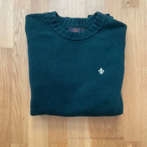 Mörkgrön stickad tröja från Morris - Mörkgrön stickad tröja från Morris med rund hals och diskret broderad logga på bröstet. Tröjan har ribbade muddar vid ärmslut och nederkant, och är perfekt för lager-på-lager. Klassisk och stilren modell som funkar till det mesta.