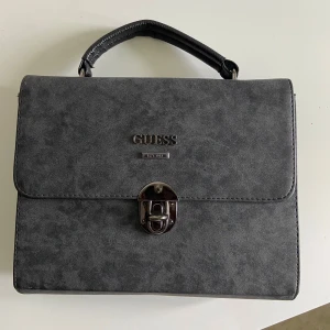 Svart handväska i mocka från Guess - Stilren svart handväska från Guess i mjuk mocka med silverfärgat lås framtill och ett smidigt handtag. Klassisk fyrkantig form och diskret Guess-logga på framsidan. Perfekt för dig som gillar en clean och tidlös look.