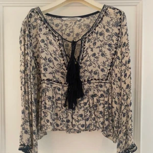 Blommig blus med snörning Zara - Supersöt blommig blus från Zara i beige och mörkblått. Lätt och luftig med långa ärmar, v-ringning och snörning med tofsar framtill. Blusen har volangdetalj nedtill och är perfekt för en chill och bohemisk vibe.