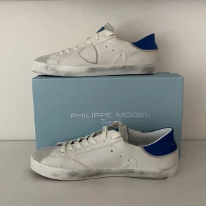 Vita Philippe Model sneakers med blå detalj - Snygga vita sneakers från Philippe Model med blå hälkappa och klassisk låg siluett. Skorna har snörning, rund tå och är tillverkade i skinn med gummisula. Perfekta för en clean och stilren look.