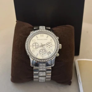 Silverfärgad Michael Kors klocka med stenar - Snygg och glittrig klocka från Michael Kors i silverfärg med stenar runt urtavlan och på länken. Urtavlan är ljus med flera små visare och datumvisning. Länken är i metall och har Michael Kors-logga på spännet. Batteriet behövs bytas