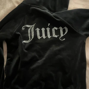 Svart velour hoodie från Juicy Couture - Säljer en svart hoodie i mjuk velour från Juicy Couture med glittrande stenar som bildar texten 'Juicy' på ryggen och JC-logga fram. Dragkedja framtill och huva. Perfekt för en trendig och avslappnad look.