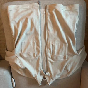 Beige overjacket med dragkedja och fickor - Säljer en beige overjacket med dragkedja framtill och två stora fickor på framsidan. Västen har en enkel och stilren design, perfekt för lager-på-lager. Passar dig som gillar minimalistisk stil. Tvättar även kläderna innan leverans!