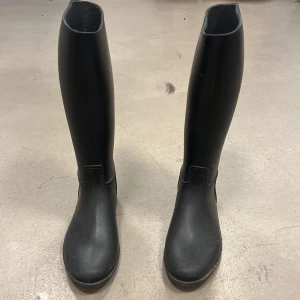 Svarta ridstövlar - Säljer ett par klassiska svarta ridstövlar i syntetmaterial. De har hög modell med rund tå och platt sula, perfekta för ridning. Enkla att matcha och praktiska för både träning och tävling.