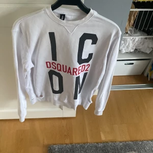 Vit sweatshirt från Dsquared2 med tryck - Vit sweatshirt från Dsquared2 med stort svart och rött 'ICON' tryck på framsidan. Tröjan har rund halsringning och långa ärmar. Klassisk passform och tillverkad i mjukt material. Man ser att den är lite använd såsom små hål ( syns på bild 2) 