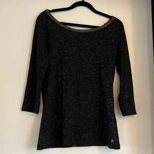 Svart glittrig klänning från Hilfiger Denim - Svart topp från Hilfiger Denim med glittriga inslag och trekvartsärm. U-ringad hals med guldfärgad kant och dekorativa knappar längs ryggen. Liten stjärndetalj nertill. Perfekt för dig som gillar stilrena men ändå festliga plagg. Passar både till vardags och till fest.