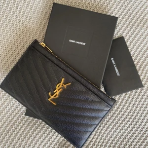 Svart kuvertväska från Saint Laurent - Säljer en svart kuvertväska från Saint Laurent med guldfärgad YSL-logga och dragkedja. Väskan har ett stilrent, quiltat mönster och är tillverkad i skinn. Köpt på NK i Stockholm, kvitto finns samt fler bilder! Nypris 3550kr. Säljer ej för längre än 2000kr! 