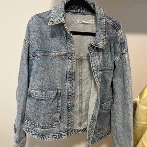 Blå jeansjacka från Zara - Säljer en klassisk blå jeansjacka från Zara i storlek S. Jackan har två stora fickor framtill, krage och knäppning med knappar. Perfekt för lager-på-lager och enkel att styla till olika outfits.