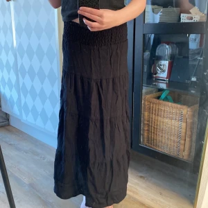 Svart långkjol från H&M - Svart långkjol från H&M med smock i midjan och lätt rynkad struktur. Kjolen har en luftig och flowy passform, perfekt för varmare dagar. Enkel och stilren design som är lätt att matcha med olika toppar.