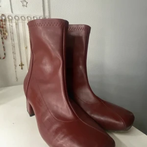 Vinröda boots med klack och dragkedja - Snygga vinröda boots i skinnimitation med fyrkantig tå och medelhög klack. Skorna har dragkedja på insidan och dekorativ söm upptill. Perfekta för att lyfta din outfit med en retrovibe.