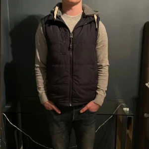 Svart dunväst med grå huva - Säljer en svart dunväst med grå huva och dragkedja framtill. Västen har fickor med knappar och en sportig look. Perfekt för lager-på-lager-stil under kyliga dagar.
