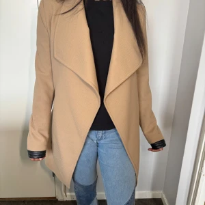 Beige vårkappa med bälte och svarta skinndetaljer - Säljer en snygg beige kappa med bälte i midjan och svarta detaljer vid ärmsluten. Jackan har en öppen, omlottliknande front och lång ärm. Perfekt för dig som vill ha en stilren och trendig look.