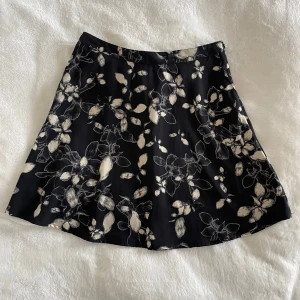 Svart blommig kjol från Åhléns - Säljer en svart kjol från Åhléns med vitt blommigt mönster. Kjolen har en lätt utsvängd form och är i ett tunt, mjukt material som känns skönt mot huden. Perfekt för dig som gillar en stilren men ändå lekfull look. Midja: 36cm, längd: 45cm