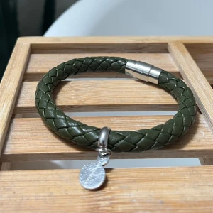 Flätat armband i grönt läder med silverdetaljer - Snyggt armband i mörkgrönt flätat läder med magnetlås i silverfärg och en rund silverberlock från Plain Vanilla.