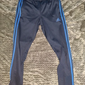 Mörkblå träningsbyxor från Adidas med blå ränder - Säljer ett par mörkblå träningsbyxor från Adidas med klassiska blå ränder längs sidorna och Adidas-logga på låret. Byxorna har elastisk midja och är tillverkade i ett mjukt, syntetiskt material. Perfekta för sport eller chill.