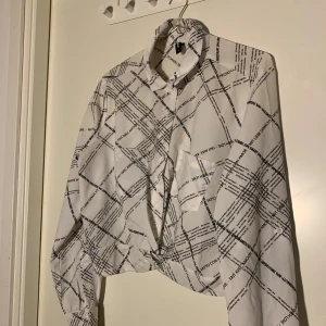 Vit croppad skjorta med svart textmönster - Säljer en vit croppad skjorta med svart grafiskt textmönster och knappar framtill. Skjortan har lång ärm och klassisk krage, perfekt för en trendig och avslappnad look.