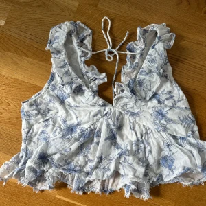Vit blommig blus med volang - Säljer en söt vit blus med blått blommönster. Blusen har volangdetaljer längs kanterna och knytning i nacken. Perfekt för sommaren.