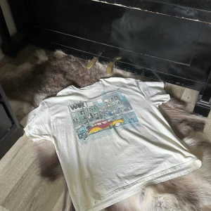 Vit oversized t-shirt med tryck från Revival Tees - Vit oversized t-shirt från Revival Tees med stort färgglatt tryck på framsidan som föreställer en stadsmiljö och en röd bil. Tillverkad i 100% bomull och har korta ärmar. Perfekt för en avslappnad stil. Tröjan är använd en gång och i nyskick. Nypris ligger på runt 800 mitt pris är endast 299