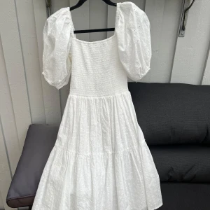 Vit klänning med puffärmar från Sisters Point - Superfin vit klänning från Sisters Point med puffiga kortärmar och smockad överdel. Klänningen har broderade detaljer och en luftig kjol som ger en romantisk känsla. Perfekt för sommarens alla tillfällen!