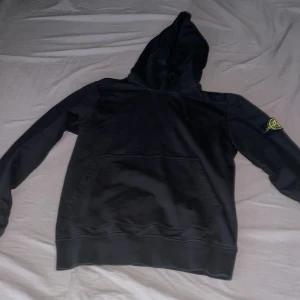 Svart hoodie från Stone Island - Säljer en svart hoodie från Stone Island med klassisk logotyp-patch på ärmen. Tröjan har huva och en stor magficka framtill. Perfekt för dig som gillar streetwear och vill ha något stilrent och bekvämt.
