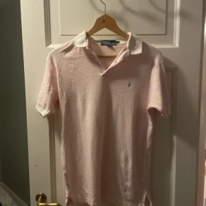Ljusrosa randig pikétröja från Polo Ralph Lauren - Den är lite missfärgad vid kragen.Säljer en ljusrosa och vit randig pikétröja från Polo Ralph Lauren. Klassisk krage i vitt och liten broderad logga på bröstet. Perfekt för en stilren och avslappnad look.