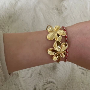 Guldfärgat armband med blommor - Säljer ett stilrent armband i guldfärg med två stora blommor som dekoration. Armbandet är öppet och justerbart, perfekt som en snygg accessoar till din outfit.