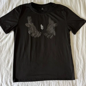 Svart t-shirt med tryck - Svart t-shirt med tryck av två händer och ett litet 'h' på bröstet. Klassisk passform och rund halsringning. Perfekt för en stilren och enkel look.