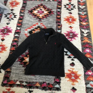 Svart stickad polotröja från Polo Ralph Lauren - Säljer en svart stickad polotröja från Polo Ralph Lauren med dragkedja vid halsen och diskret logga på bröstet. Tröjan har lång ärm och ribbade muddar. Perfekt för en stilren och klassisk look.