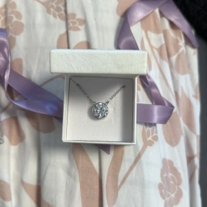 Silverfärgat halsband med gnistrande sten - Säljer ett elegant halsband i silverfärg med en rund, glittrig sten i mitten. Kedjan är tunn och smycket levereras i en vit ask. Perfekt för att ge en extra touch till din outfit. Oanvänd 