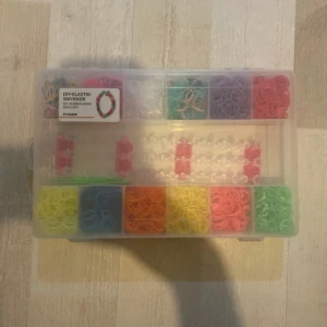 DIY elastikarmbandskit från Tiger - Säljer ett komplett DIY-kit för att göra egna elastiska armband. Kitet innehåller färgglada gummiband i flera färger, två gröna krokar, plastclips och en armbandsväv. Perfekt för att skapa personliga smycken!