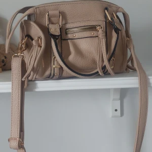 Beige axelväska med gulddetaljer - Säljer en beige axelväska med guldfärgade metalldetaljer och svarta kanter. Väskan har både handtag och justerbar axelrem, samt flera fack med dragkedja. Insidan är fodrad med randigt tyg. Perfekt för att bära dina viktigaste saker.