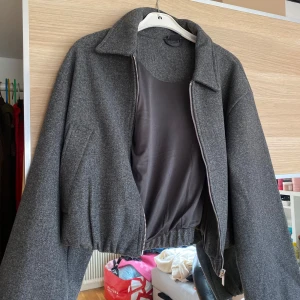 Mörkgrå cropped jacka med dragkedja - Säljer en mörkgrå jacka med croppad passform och dragkedja framtill. Jackan har lång ärm, klassisk krage och resår i midjan för en snygg siluett. Perfekt för dig som gillar stilrena och trendiga plagg.