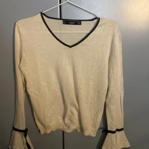 Beige långärmad topp  - Säljer en beige långärmad topp från Mango med svarta detaljer vid v-ringning och ärmslut. Toppen har volangdetaljer vid ärmslut och en mjuk, tunn känsla. 