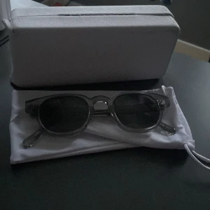 Chimi 01 - Snygga solglasögon med transparent grå båge och runda, mörka glas. Kommer med ett vitt fodral och hårt etui. Perfekta för soliga dagar och ger en cool look. 