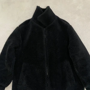 Svart teddyjacka från Bershka - Mysig svart teddyjacka från Bershka med hög krage. Jackan har två fickor och är perfekt för kyliga dagar. Den fluffiga ytan ger en trendig look och passar bra till många outfits.