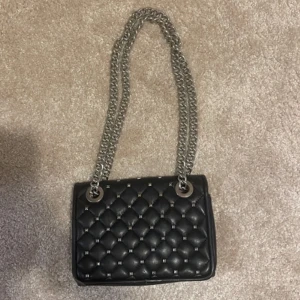 Svart quiltad axelväska med kedja - Säljer en svart quiltad axelväska med silverfärgad kedjerem och metallnitar. Väskan har magnetlås och är tillverkad i skinnimitation. Perfekt storlek för det viktigaste och ger en cool touch till din outfit.