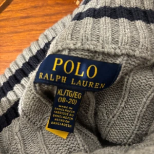 Grå kabelstickad tröja från Polo Ralph Lauren - Säljer en klassisk grå kabelstickad tröja från Polo Ralph Lauren. Tröjan har lång ärm, rund halsringning och en liten broderad logga på bröstet. Perfekt för en stilren och tidlös look.