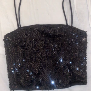 Svart paljett-tubtopp från Lindex - Säljer en svart tubtopp från Lindex med tunna axelband och täckt av glittriga paljetter. Perfekt för att sticka ut och ge outfiten extra bling. Toppen är croppad och har en figurnära passform. Bara testad