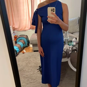 Kornblå one shoulder långklänning med rosett - Säljer en kornblå långklänning med one shoulder-design och stor rosett på axeln. Klänningen har en tight passform och är tillverkad i ett ribbat material som ger en snygg siluett. Perfekt för dig som vill sticka ut med en elegant och modern look.