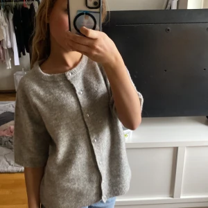 Grå stickad kofta från 157 - Mysig grå stickad kofta från 157 med pärlknappar och korta ärmar. Koftan har rund halsringning och ribbade kanter. Perfekt att slänga över en topp för en enkel och stilren look.