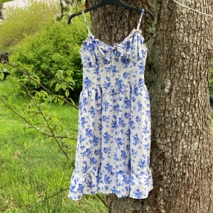 Blommig sommarklänning med volang - Söt vit klänning med blått blommönster och volangdetalj nertill. Axelbandsmodell med smockad överdel och knyt framtill. Perfekt för varma dagar och midsommarfirande.